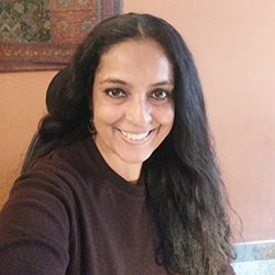 Anubha Kathuria Bellani PCC headshot photo, dark haired woman wearing dark top in front orange wall smiling