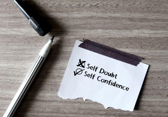 pen and marker next to piece of paper with an x next to self doubt and a check mark next to self confidence