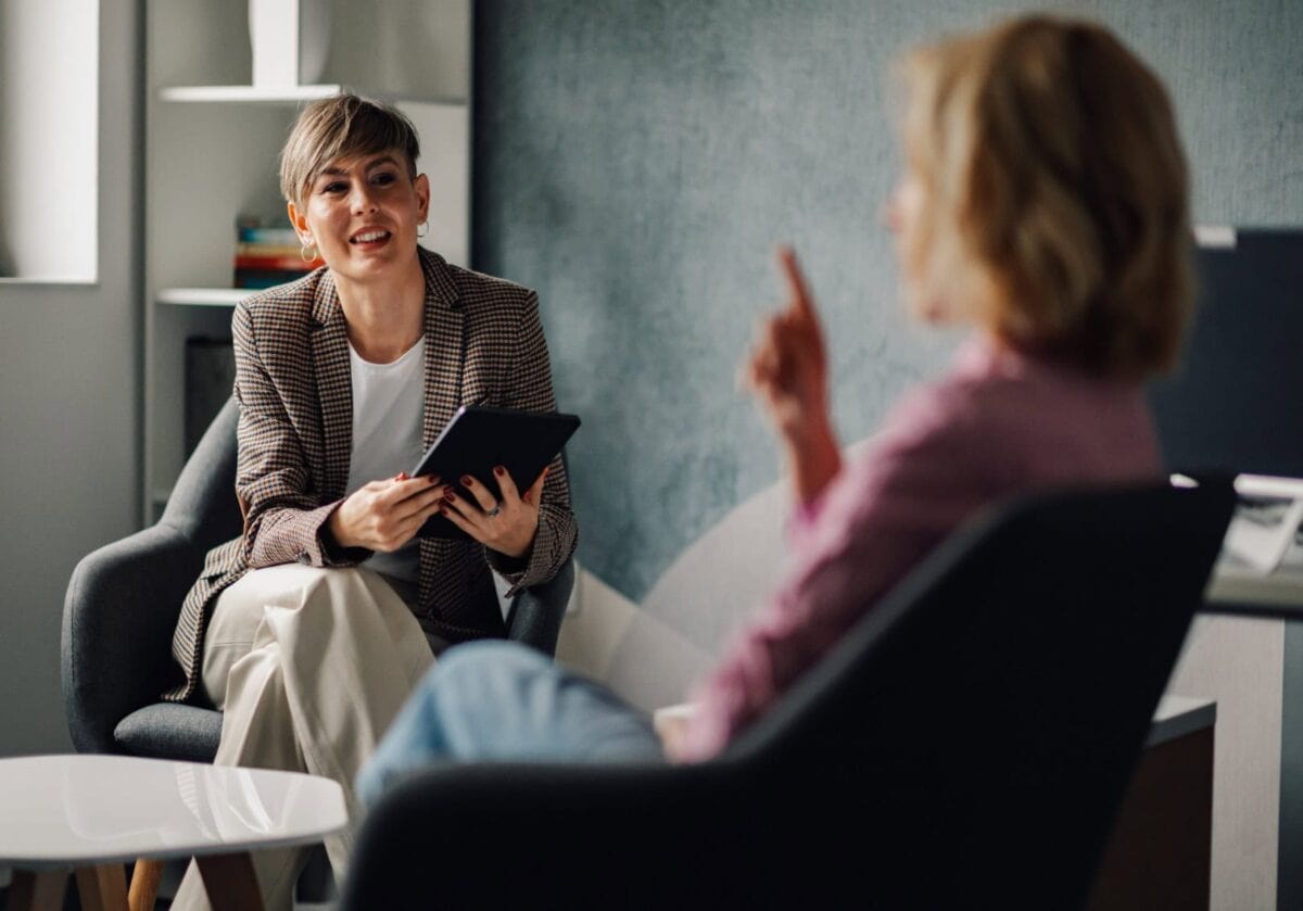 A female coach smiling while holding a tablet and listening to a client seated opposite her in a relaxed office setting.