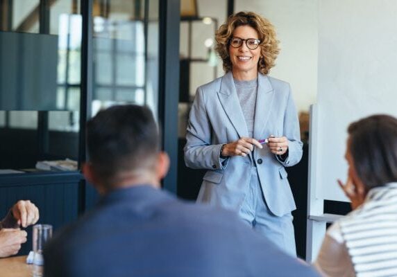 woman in a conference room looking confident and speaking to a group of business professionals seated at the table