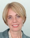 Meg Moore headshot photo, smiling woman with blond hair and gray blazer in front of white wall