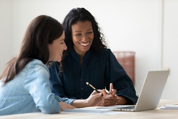 two women sitting at a laptop taking notes and engaging in conversation